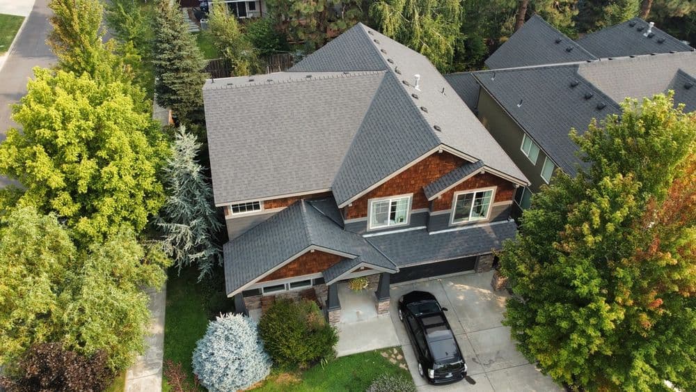 Aerial view of a modern home with a dark roof surrounded by lush green trees and a driveway.