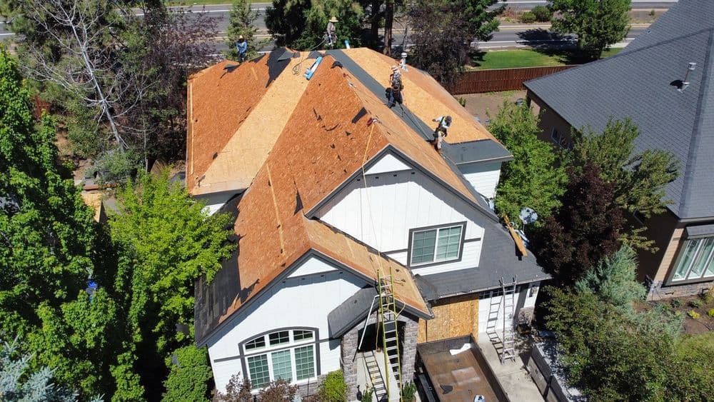 Workers installing new roofing on a residential house with a tree-lined background.