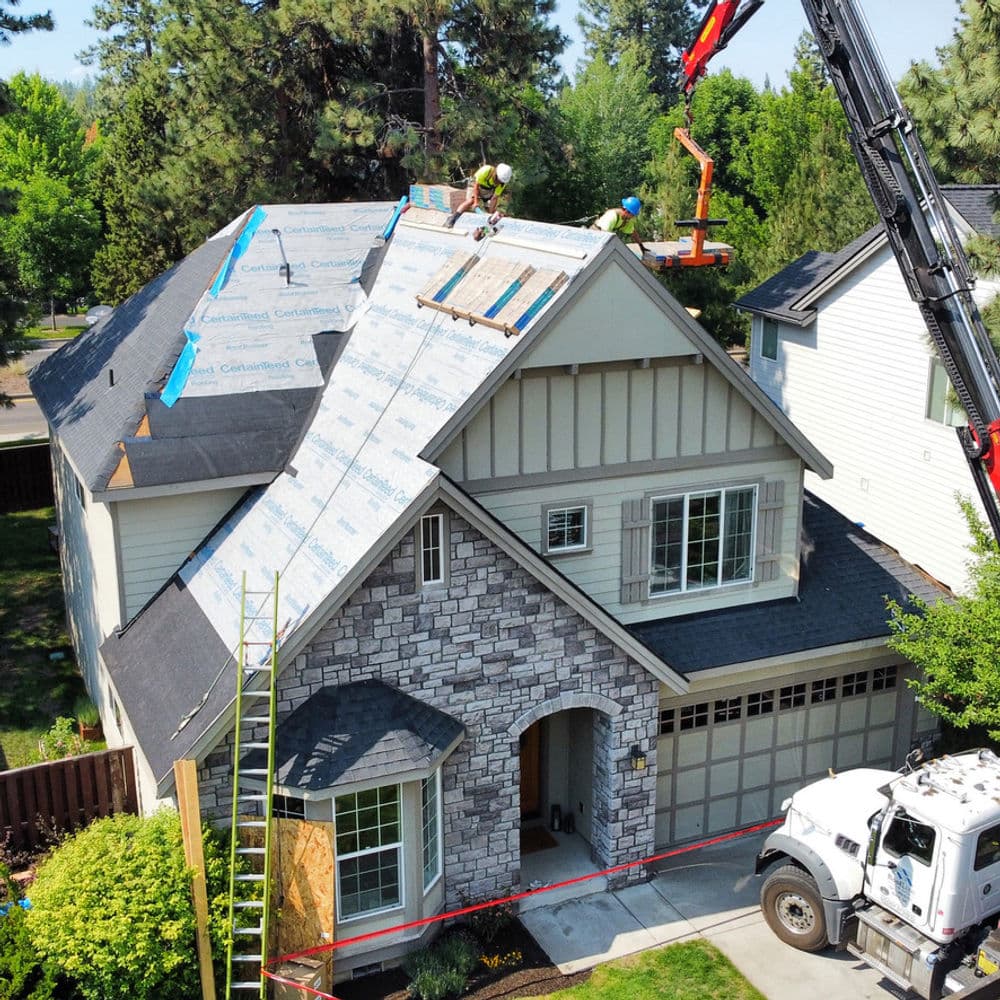 Roofing contractors working on a residential home with a crane and materials on the roof.