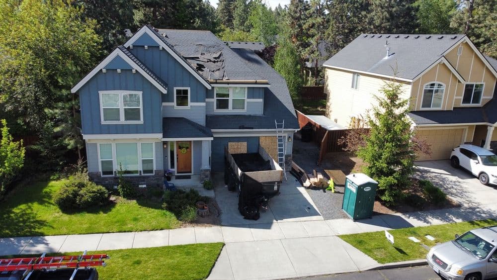 Aerial view of a blue two-story house under roof repair with a dumpster and portable toilet outside.